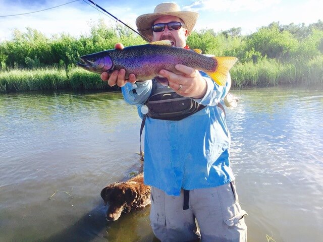 Attorney Jack Edwards holding beautiful just caught rainbow trout while standing in the water with his chocolate lab dog